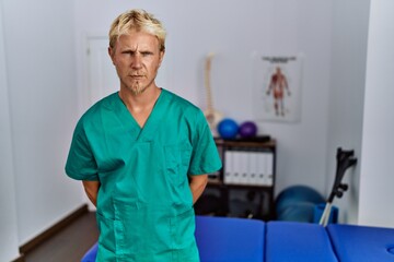 Young blond man wearing physiotherapist uniform standing at clinic skeptic and nervous, frowning upset because of problem. negative person.