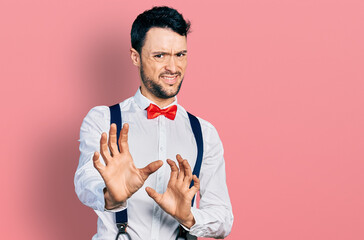 Hispanic man with beard wearing hipster look with bow tie and suspenders disgusted expression, displeased and fearful doing disgust face because aversion reaction. with hands raised