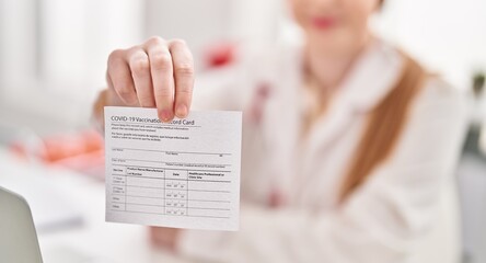 Young blonde woman wearing doctor uniform hoding covid-19 vaccination record card at clinic