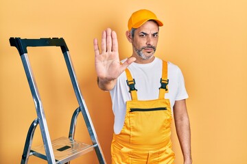 Handsome middle age man with grey hair holding ladder doing stop sing with palm of the hand. warning expression with negative and serious gesture on the face.