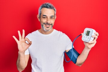 Handsome middle age man with grey hair using blood pressure monitor doing ok sign with fingers,...
