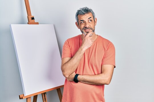 Handsome middle age man with grey hair standing by painter easel stand thinking worried about a question, concerned and nervous with hand on chin