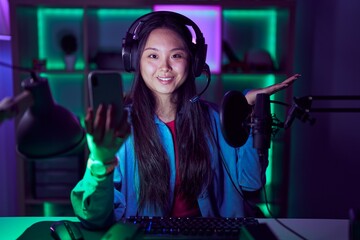 Young asian woman playing video games with smartphone smiling cheerful presenting and pointing with palm of hand looking at the camera.