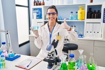 Hispanic woman working at scientist laboratory smiling happy and positive, thumb up doing excellent and approval sign