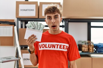 Young caucasian volunteer man holding envelope with money at donations stand scared and amazed with...