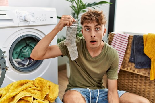 Young caucasian man doing laundry holding sock scared and amazed with open mouth for surprise, disbelief face
