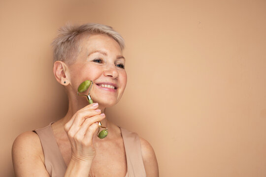 Gorgeous woman of 60s massaging face skin using natural facial stone trendy jade green quartz roller