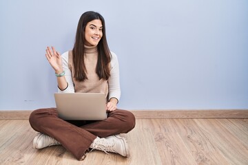 Young brunette woman working using computer laptop sitting on the floor waiving saying hello happy and smiling, friendly welcome gesture