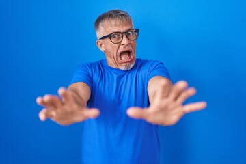 Hispanic man with grey hair standing over blue background afraid and terrified with fear expression stop gesture with hands, shouting in shock. panic concept.