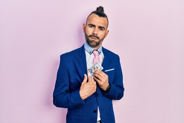 Young hispanic man wearing business suit holding dollar clueless and confused expression. doubt...