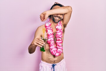 Young hispanic man wearing swimsuit and hawaiian lei drinking tropical cocktail smiling cheerful...