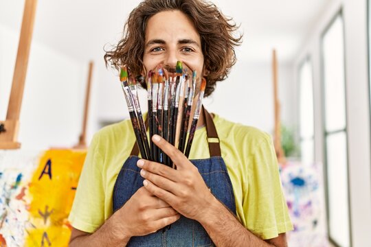 Young hispanic artist man smiling happy holding paintbrushes at art studio.