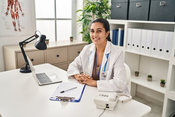 Young hispanic woman wearing doctor uniform looking to the camera at clinic