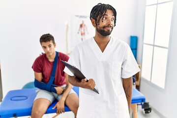 Obraz premium Young hispanic man working at pain recovery clinic with a man with broken arm smiling looking to the side and staring away thinking.