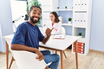Obraz premium Young hispanic man at doctor clinic pointing thumb up to the side smiling happy with open mouth