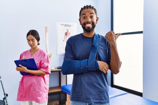 Young hispanic man wearing arm on sling at physiotherapy clinic pointing thumb up to the side smiling happy with open mouth
