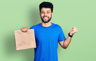 Young arab man with beard holding take away paper bag screaming proud, celebrating victory and...