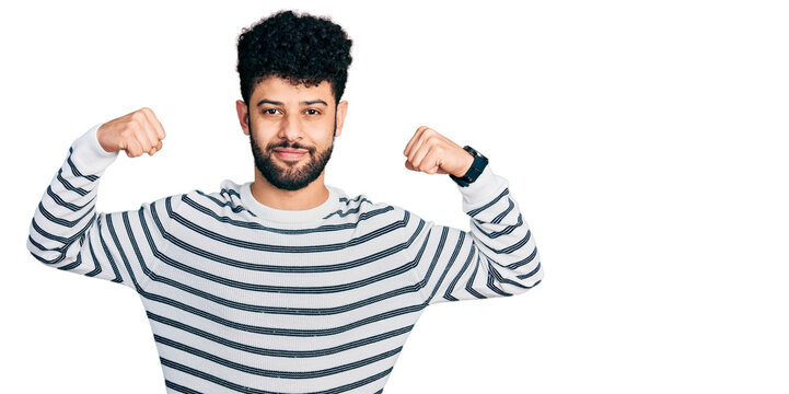 Young arab man with beard wearing casual striped sweater showing arms muscles smiling proud. fitness concept.