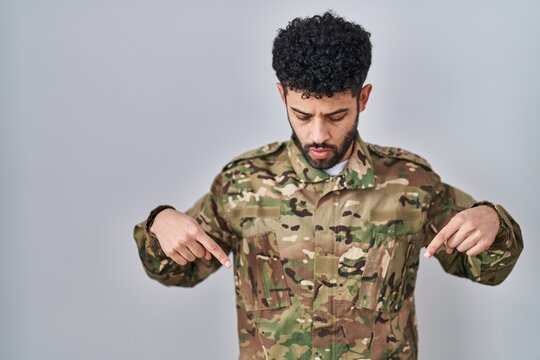Arab man wearing camouflage army uniform pointing down with fingers showing advertisement, surprised face and open mouth