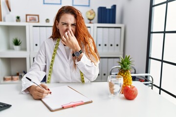 Young redhead woman nutritionist doctor at the clinic smelling something stinky and disgusting,...