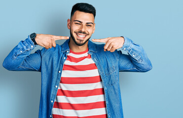 Young hispanic man with beard wearing casual denim jacket smiling cheerful showing and pointing...