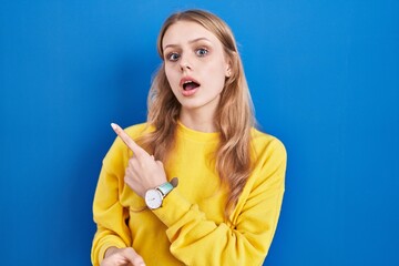 Young caucasian woman standing over blue background surprised pointing with finger to the side, open mouth amazed expression.