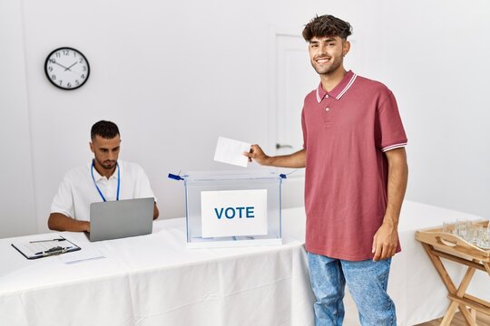 Young hispanic voter man smiling happy putting vote in ballot box at electoral center.