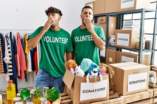 Young gay couple wearing volunteer t shirt at donations stand bored yawning tired covering mouth with hand. restless and sleepiness.