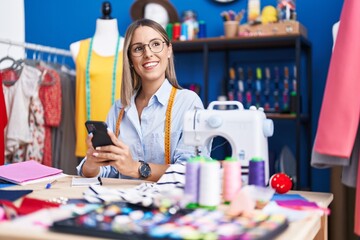 Young beautiful hispanic woman tailor smiling confident using smartphone at clothing factory