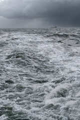 Vertical shot of very rough sea with black dramatic clouds in the sky and small ship on the horizon. Storm at the sea. Stormy sea with big waves. North sea close to shore of Germany