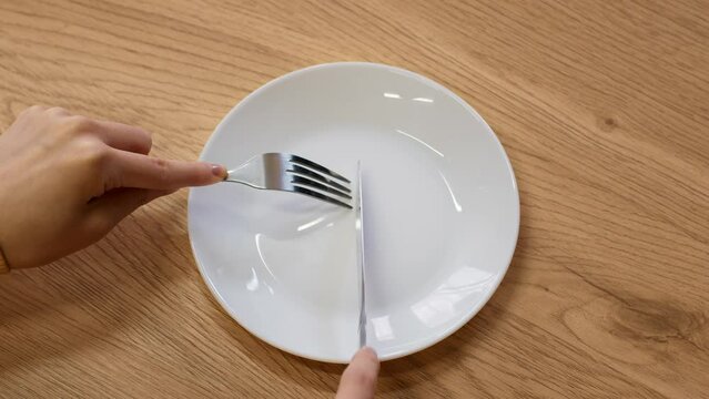 Hunger crisis. Top view of female hands with knife and fork cutting invisible food in empty clean plate, slow motion