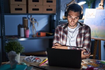 Young hispanic man sitting at art studio with laptop late at night skeptic and nervous, disapproving expression on face with crossed arms. negative person.