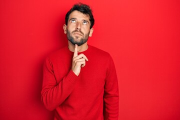 Handsome man with beard wearing casual red sweater thinking concentrated about doubt with finger on chin and looking up wondering
