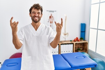Young handsome physiotherapist man working at pain recovery clinic shouting with crazy expression doing rock symbol with hands up. music star. heavy music concept.