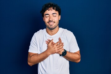 Young hispanic man wearing casual white t shirt smiling with hands on chest with closed eyes and grateful gesture on face. health concept.