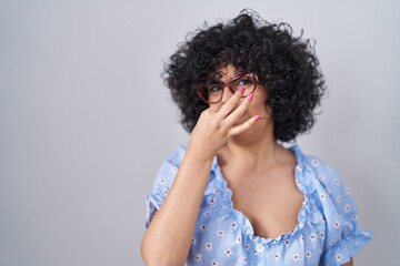 Young brunette woman with curly hair wearing glasses over isolated background smelling something stinky and disgusting, intolerable smell, holding breath with fingers on nose. bad smell