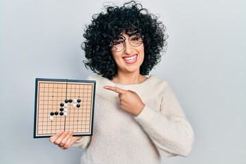 Young middle east woman holding asian go game board smiling happy pointing with hand and finger