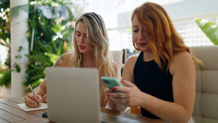 Two women using smartphone writing on notebook sitting on table at home terrace