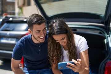 Fototapeta na wymiar Young hispanic couple sitting on car trunk watching video on smartphone at street