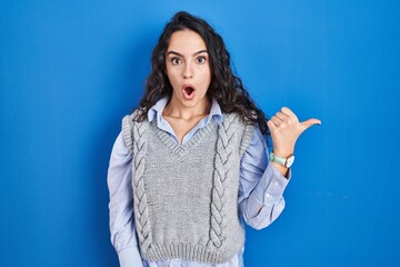 Young brunette woman standing over blue background surprised pointing with hand finger to the side, open mouth amazed expression.