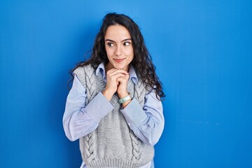Young brunette woman standing over blue background laughing nervous and excited with hands on chin...