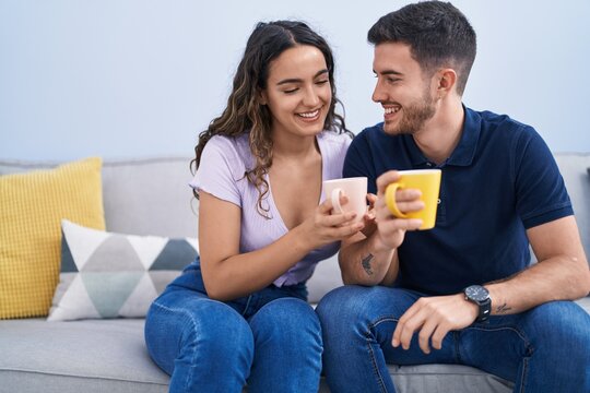 Young hispanic couple drinking coffee sitting on sofa at home