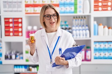 Young caucasian woman working at pharmacy drugstore holding pills clueless and confused expression....