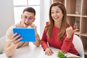 Mother and son using touchpad device pointing thumb up to the side smiling happy with open mouth