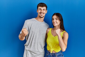 Fototapeta premium Young hispanic couple standing together over blue background very happy and excited doing winner gesture with arms raised, smiling and screaming for success. celebration concept.