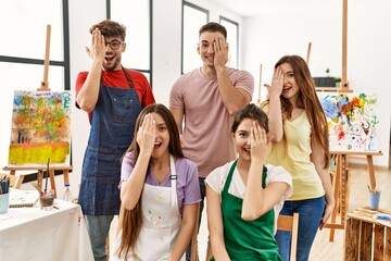 Group of five hispanic artists at art studio covering one eye with hand, confident smile on face and surprise emotion.