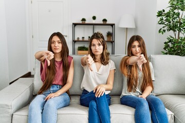 Group of three hispanic girls sitting on the sofa at home looking unhappy and angry showing rejection and negative with thumbs down gesture. bad expression.