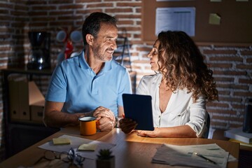 Middle age hispanic couple using touchpad sitting on the table at night looking to side, relax profile pose with natural face and confident smile.