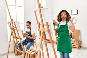 Hispanic middle age woman and mature man at art studio celebrating victory with happy smile and winner expression with raised hands