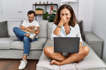 Hispanic middle age couple at home, woman using laptop smelling something stinky and disgusting, intolerable smell, holding breath with fingers on nose. bad smell - obrazy, fototapety, plakaty